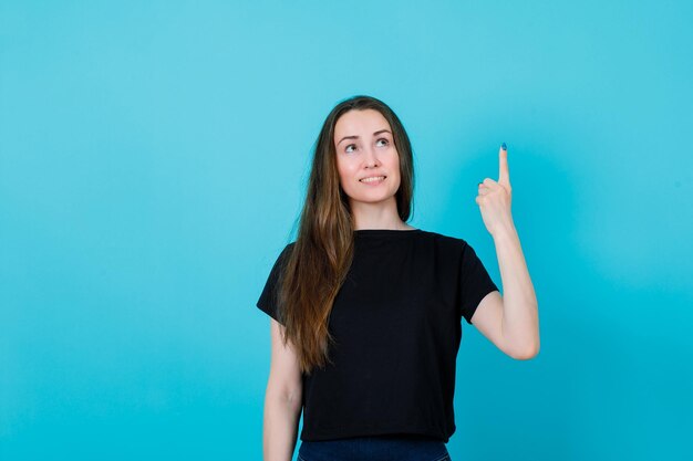 Nach oben schauendes Mädchen zeigt mit dem Zeigefinger auf blauem Hintergrund nach oben