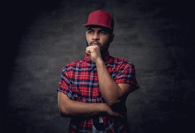 Nachdenklich ein afroamerikanischer Hipster-Typ, gekleidet in ein rotes Fleece-Shirt und eine Mütze im Studio. Getrennt auf einem dunklen strukturierten Hintergrund.