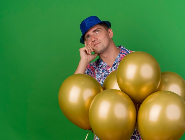 nachdenklich junger Party-Typ, der blauen Hut trägt, der hinter Luftballons steht und Hand auf Wange lokalisiert auf Grün setzt