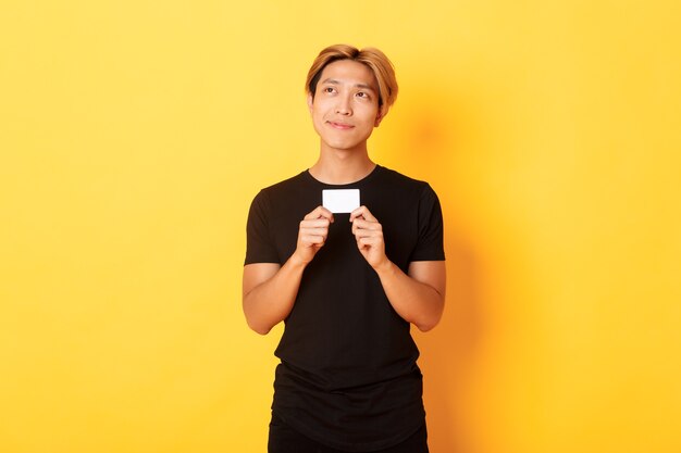 Nachdenklich lächelnder asiatischer Typ, der denkt, während er Kreditkarte zeigt und verträumte gelbe Wand der oberen linken Ecke schaut