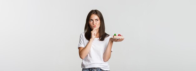 Nachdenklich lächelndes hübsches Mädchen, das nachdenkt, während es ein Stück Kuchen auf weißem Hintergrund hält