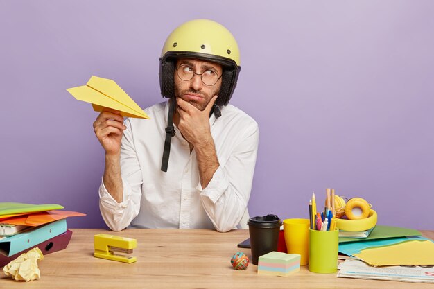 Nachdenklich nachdenklich junger Mann müde von der Arbeit im Büro, hält Papier handgemachtes Flugzeug, trägt Schutzhelm, weißes Hemd, hält Kinn, denkt über den Wechsel der Arbeitsposition