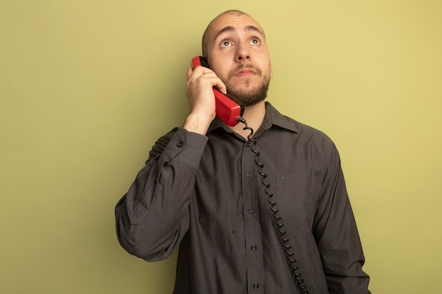 nachdenklich nachschlagen junger hübscher Kerl, der schwarzes T-Shirt trägt, spricht am Telefon isoliert auf Olivgrün