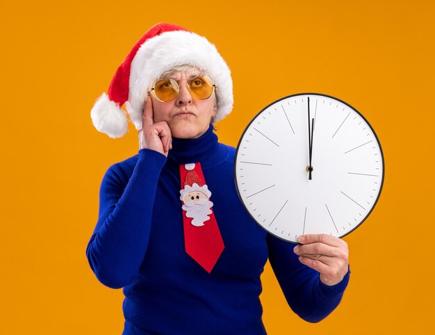 Nachdenkliche ältere Frau in Sonnenbrille mit Weihnachtsmütze und Weihnachtsmann-Krawatte legt den Finger auf den Tempel und hält die Uhr isoliert an der orangefarbenen Wand mit Kopierraum