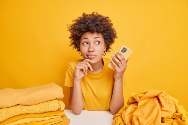 Nachdenkliche Afro-Amerikanerin hat verträumten Ausdruck hält modernes Handy sitzt am Tisch mit Haufen von Kleidung isoliert über gelber Wand beschäftigt Wäsche falten. Kleidung und Haushalt