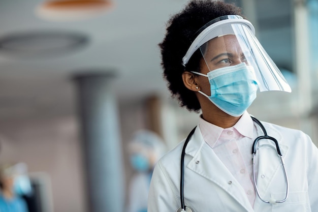 Nachdenkliche afroamerikanische Ärztin mit Gesichtsschutz und Maske im Krankenhaus