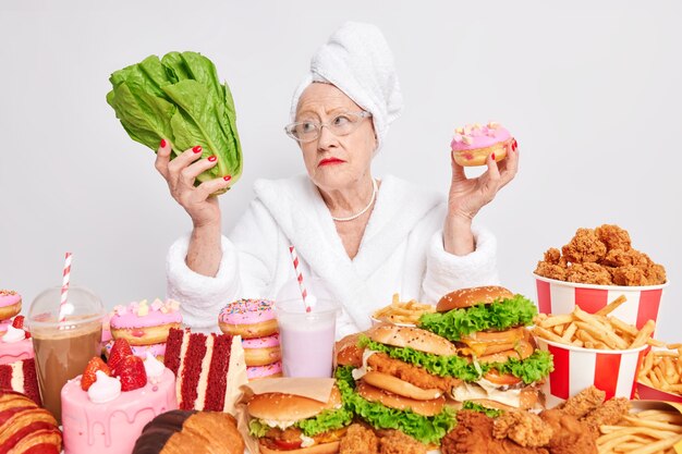 Nachdenkliche alte Frau wählt zwischen gesundem und ungesundem Essen hält grünen Salat und leckeren leckeren Donut