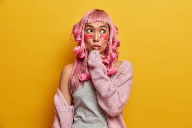 Nachdenkliche asiatische Frau mit rosa Haaren, bereitet sich auf besondere Anlässe vor, trägt Kollagenpads und Lockenwickler auf, berührt die Lippen, konzentriert darüber