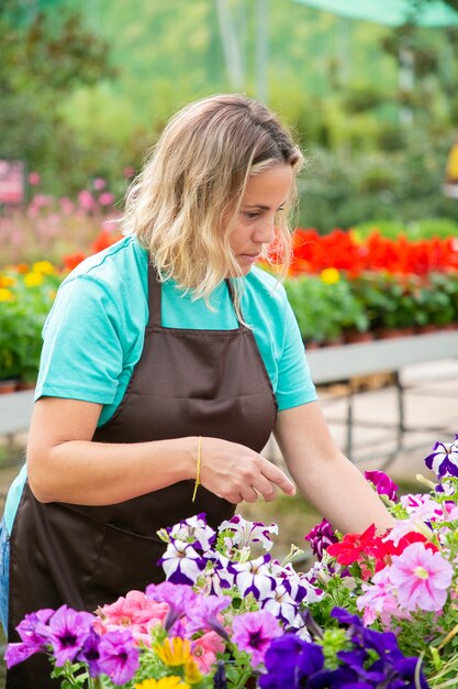 Nachdenkliche blonde Frau, die Blumenpflanzen in Töpfen betrachtet