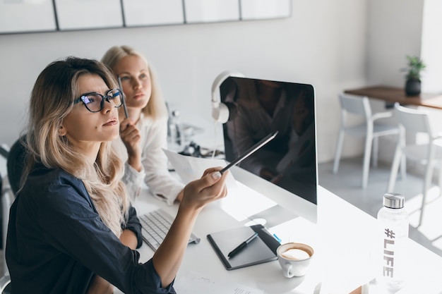 Nachdenkliche blonde Frau in den Gläsern, die Bleistift halten und weg während der Arbeit im Büro schauen. Innenporträt der beschäftigten langhaarigen Buchhalterin unter Verwendung des Computers.