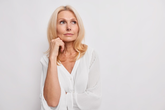 Nachdenkliche blonde Frau mittleren Alters denkt über etwas nach, hält die Hand in der Nähe des Gesichts hat gesunde Haut minimales Make-up trifft die Wahl trägt weiße Bluse posiert im Innenbereich für leere Kopien für Ihre Werbung