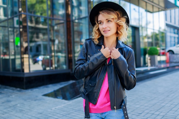 Nachdenkliche blonde kurzhaarige Frau, die auf Straßen der großen modernen Stadt geht. Modisches urbanes Outfit. Ungewöhnliche rosa Sonnenbrille.