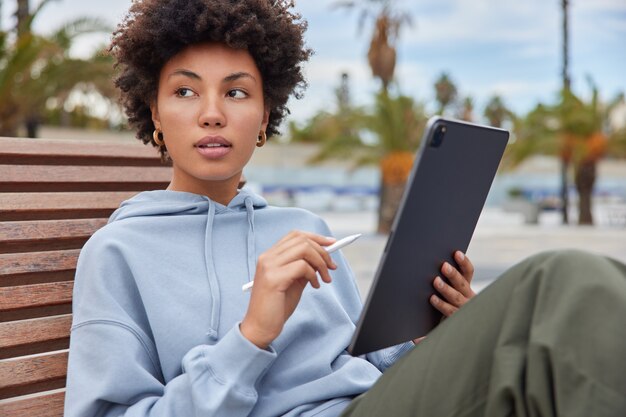 Nachdenkliche Designerin hält Tablet mit Stift und verwendet eine öffentliche Internetverbindung zum Erstellen von Medienskizzen über eine Anwendung für kreative Künstler, die lässig gekleidet auf Holzbankposen draußen posieren