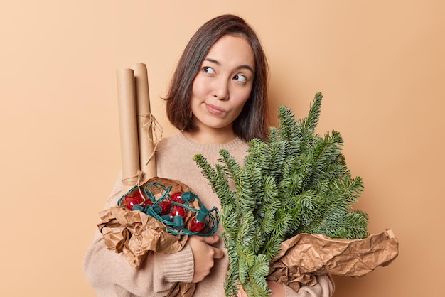 Nachdenkliche dunkelhaarige Frau, die konzentriert wegblickt, mit verträumtem Gesichtsausdruck, hält Fichtenzweige, die in eine Retro-Girlande aus Papier gewickelt sind, bereitet sich auf Winterferien vor, isoliert auf beigem Hintergrund