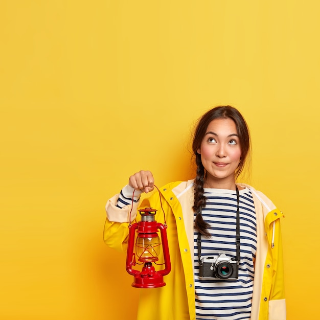 Nachdenkliche dunkelhaarige Frau mit gekämmtem Zopf, hält Petroleumlampe, gekleidet in gestreiften Pullover, Regenmantel, verwendet Retro-Kamera zum Fotografieren, schaut nachdenklich nach oben, denkt über die nächste Reise nach