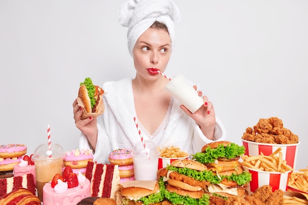 Nachdenkliche europäische Dame mit rot geschminkten Lippen trinkt Limonade isst Burger sieht weg süchtig nach Fast Food