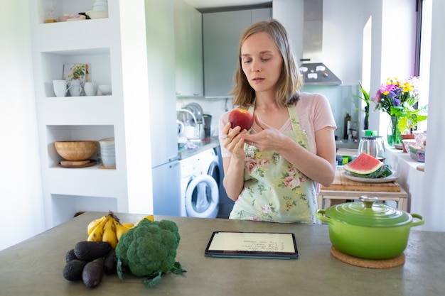 Nachdenkliche fokussierte Frau, die Obst beim Kochen in der Küche hält, mit Tablette nahe Topf und frischem Gemüse auf Theke. Vorderansicht. Kochen zu Hause und gesundes Essen Konzept