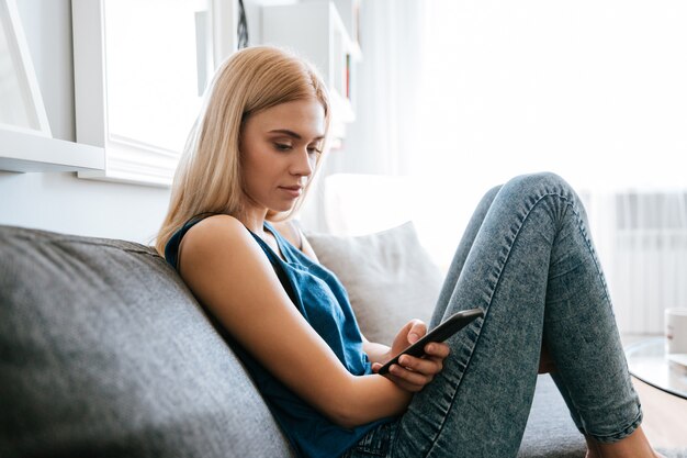 Nachdenkliche Frau, die auf Sofa sitzt und Handy zu Hause benutzt