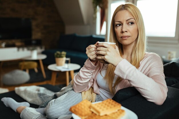 Nachdenkliche Frau, die Kaffee trinkt, während sie auf dem Sofa sitzt und zu Hause an etwas denkt