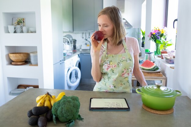 Nachdenkliche Frau, die Obst beim Kochen in ihrer Küche riecht, mit Tablette nahe Topf und frischem Gemüse auf Theke. Vorderansicht. Kochen zu Hause und gesundes Essen Konzept