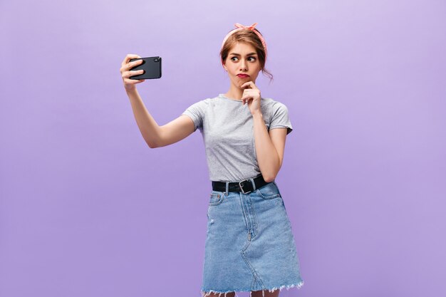 Nachdenkliche Frau im Jeansrock nimmt Selfie auf lila Hintergrund. Junges Mädchen mit rosa Kopftuch im grauen T-Shirt macht Foto.