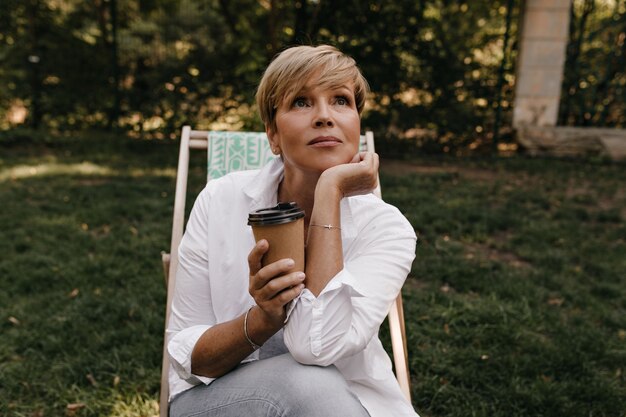 Nachdenkliche Frau mit stilvoller blonder Frisur in der weißen kühlen Bluse und in den Jeans, die Tasse Kaffee halten und im Park nachschlagen.