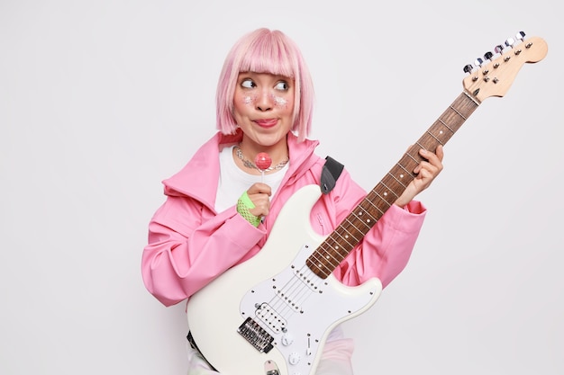 Nachdenkliche Frau mit trendiger rosa Frisur leckt die Lippen hält süße Lollipop-Posen im Innenbereich hält akustische Bass-E-Gitarre in stilvollem Outfit