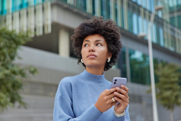 Nachdenkliche Frau ruht im Freien und konzentriert sich über Blogs in sozialen Netzwerken über Mobilfunk, die mit Highspeed-Internet verbunden sind, genießt gutes Wetter und trägt einen lässigen blauen Pullover Moderne Technologien und Lifestyle