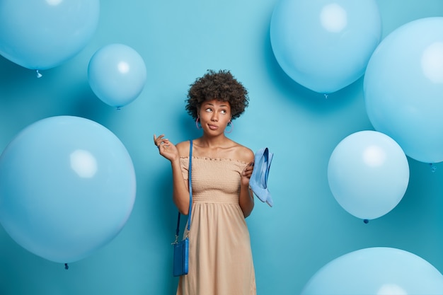 Nachdenkliche Frau trägt langes beiges Kleid, hält blaue Schuhe mit hohen Absätzen passend zur Tasche, kommt zum Jubiläum eines Freundes, bereit für ein festliches Ereignis, isoliert über blauer Wand mit aufgeblasenen Luftballons