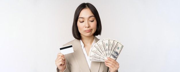 Nachdenkliche Geschäftsfrau koreanische Unternehmensfrau, die Kreditkarte und Geldbargelddollar in den Händen zeigt, die über weißem Hintergrund stehen und denken