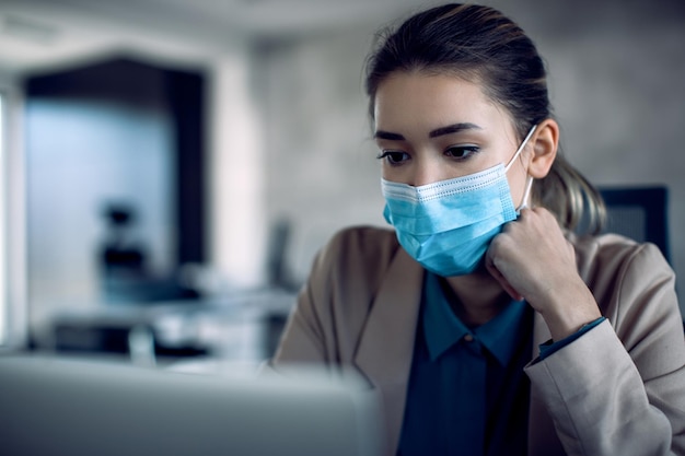 Nachdenkliche Geschäftsfrau mit Gesichtsmaske, die eine E-Mail auf einem Computer im Büro liest