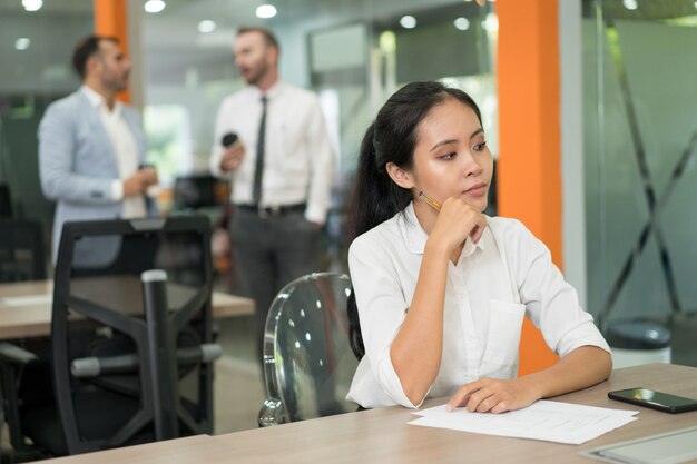 Nachdenkliche hübsche asiatische Geschäftsfrau, die am Schreibtisch im Büro sitzt