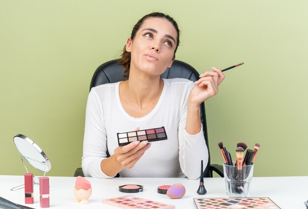 Nachdenkliche hübsche kaukasische Frau, die am Tisch mit Make-up-Tools sitzt und Lidschatten-Palette und Make-up-Pinsel zur Seite hält