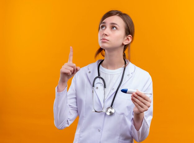 Nachdenkliche junge Ärztin, die medizinische Robe und Stethoskop trägt und Thermometer mit erhobenem Finger auf isolierter orange Wand mit Kopienraum hält