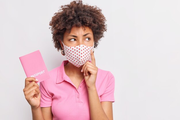 Nachdenkliche junge Afro-Amerikanerin denkt darüber nach, wo sie Urlaub machen soll, Reisepass trägt schützende Gesichtsmaske in rosafarbenem T-Shirt isoliert über weißer Wand