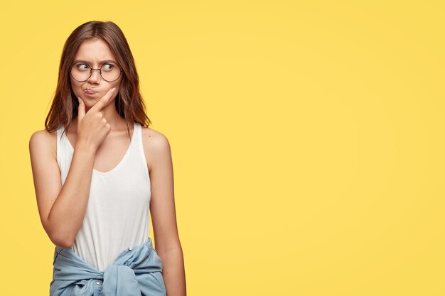 Nachdenkliche junge Brünette mit Brille, die gegen die gelbe Wand aufwirft