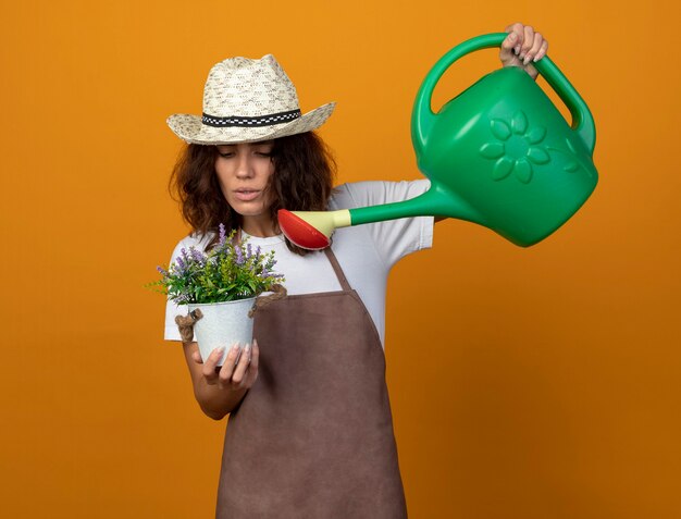 nachdenkliche junge Frau Gärtnerin in Uniform mit Gartenhut, die Blume im Blumentopf mit Gießkanne tränkt, isoliert auf Orange