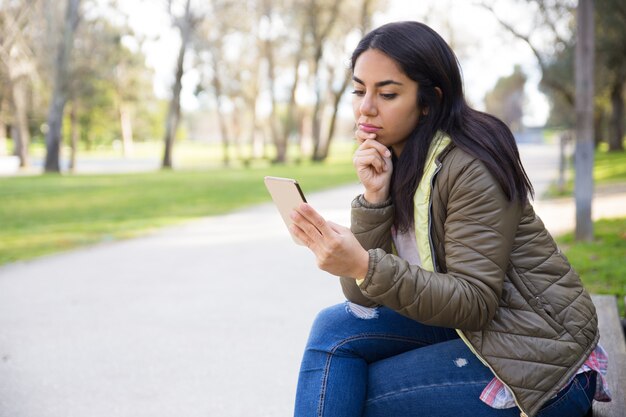 Nachdenkliche junge Frau im surfenden Netz der Jacke auf Smartphone