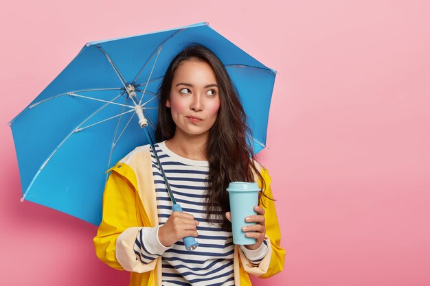 Nachdenkliche junge Frau mit asiatischem Aussehen, geht während des regnerischen bewölkten Tages unter Regenschirm, trinkt Kaffee zum Mitnehmen