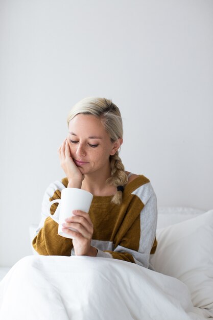 Nachdenkliche junge Frau sitzt auf dem Bett mit Tasse