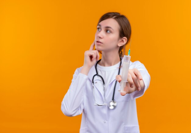 Nachdenkliche junge Ärztin, die medizinische Robe und Stethoskop-Streckspritze trägt, die linke Seite auf isolierter orange Wand mit Kopienraum betrachten