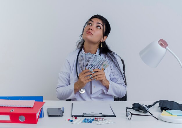 Nachdenkliche junge Ärztin, die medizinische Robe und Stethoskop trägt, sitzt am Schreibtisch mit medizinischen Werkzeugen, die Geld halten, das lokal nach oben schaut