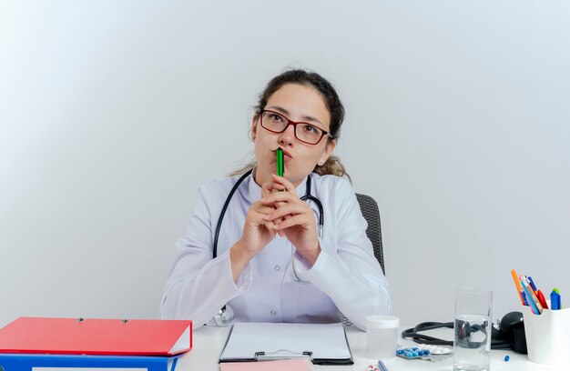 Nachdenkliche junge Ärztin, die medizinische Robe und Stethoskop und Brille trägt, sitzt am Schreibtisch mit medizinischen Werkzeugen, die Lippen mit Stift suchen, der lokal nach oben schaut