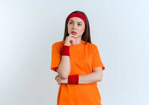 Nachdenkliche junge sportliche Frau, die Stirnband und Armbänder trägt, die Kinn berühren, betrachten Seite lokalisiert