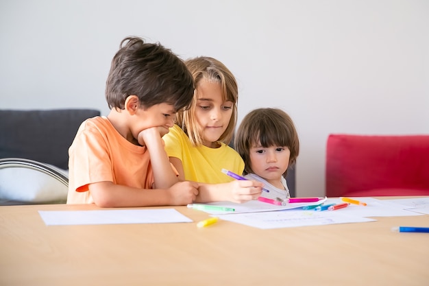 Nachdenkliche Kinder malen mit Markern im Wohnzimmer. Drei kaukasische entzückende Kinder, die zusammen sitzen, das Leben genießen, zeichnen und zusammen spielen. Kindheit, Kreativität und Wochenendkonzept