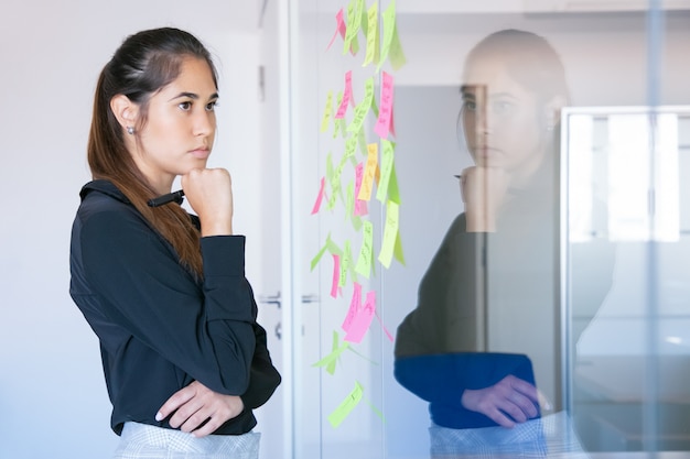 Nachdenkliche lateinische Geschäftsfrau, die Markierung hält und Notizen auf Glaswand liest. Konzentrierte sich zuversichtlich, hübsche Arbeiterin im Anzug, die über die Idee für ein Projekt nachdenkt.