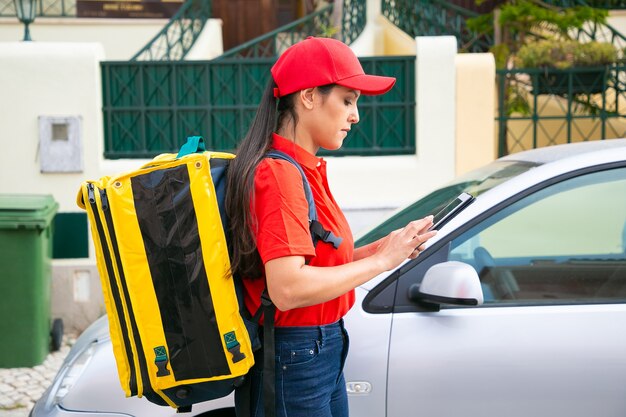 Nachdenkliche Lieferfrau beobachtet erforderliches Haus auf Tablette. Junger weiblicher Kurier mit gelbem Thermorucksack, der Expressauftrag liefert und auf Straße geht. Lieferservice und Online-Shopping-Konzept