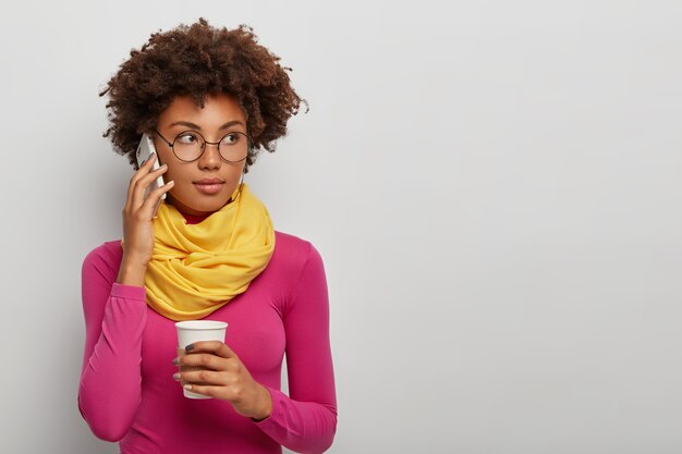 Nachdenkliche lockige Frau hat Telefongespräch über Handy, trinkt Kaffee, genießt Unterhaltung, trägt eine Brille, rosa Rollkragenpullover mit gelbem Schal, posiert vor weißem Hintergrund
