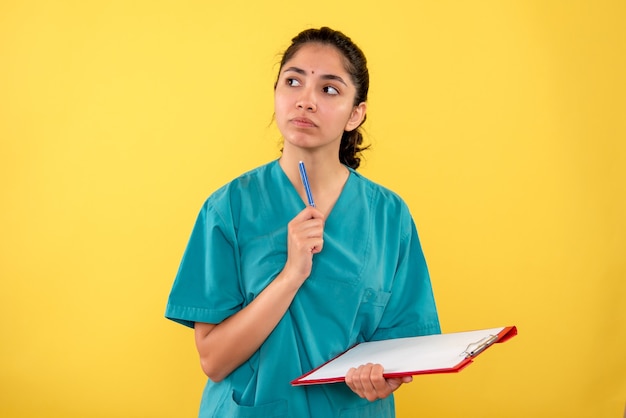 Nachdenkliche nachdenkliche Ärztin in Uniform, die Zwischenablage und Stift auf gelbem Hintergrund hält