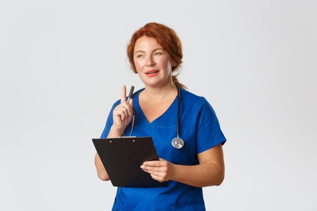 Nachdenkliche rothaarige Ärztin, rothaarige Ärztin in blauen Peelings, die vom Patientenfall fasziniert ist, Stift schüttelt und Zwischenablage hält